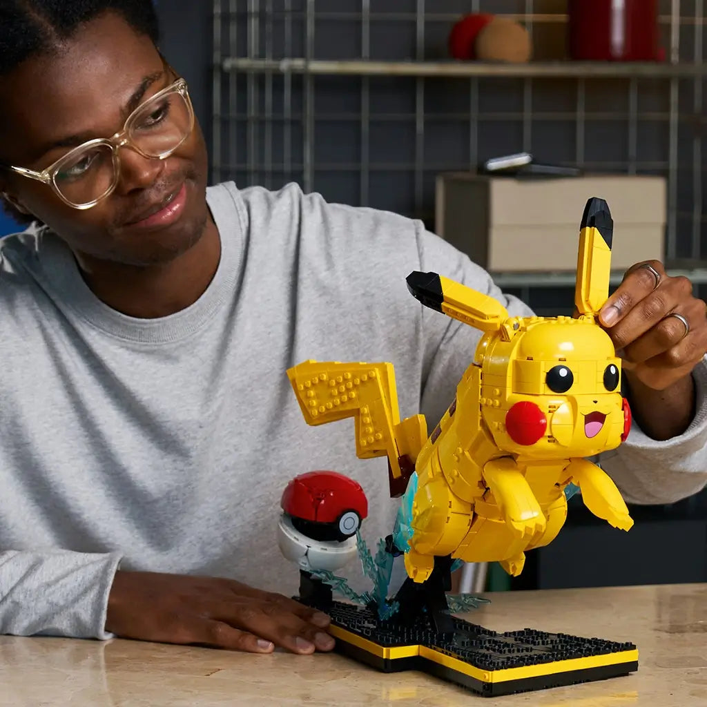 LEGO Pokémon Pikachu model features bright yellow plastic construction, a rounded character form with black-tipped ears, displayed with a Poké Ball while a builder adjusts the figure on a tabletop setting.