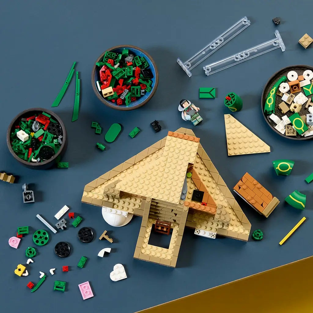 LEGO Pokémon Rayquaza building set is partly assembled in tan, green, red, and white bricks, with loose pieces, clear supports, and a minifigure spread on a blue tabletop.