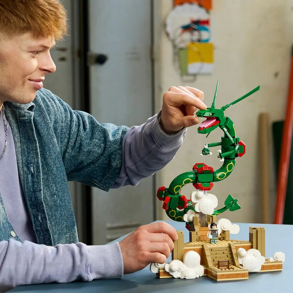 LEGO Pokémon Rayquaza set in green, red, tan, and white bricks is adjusted by an adult builder, with the articulated dragon rising above a cloud-covered shrine on a table.