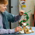 LEGO Pokémon Rayquaza set in green, red, tan, and white bricks is adjusted by an adult builder, with the articulated dragon rising above a cloud-covered shrine on a table.