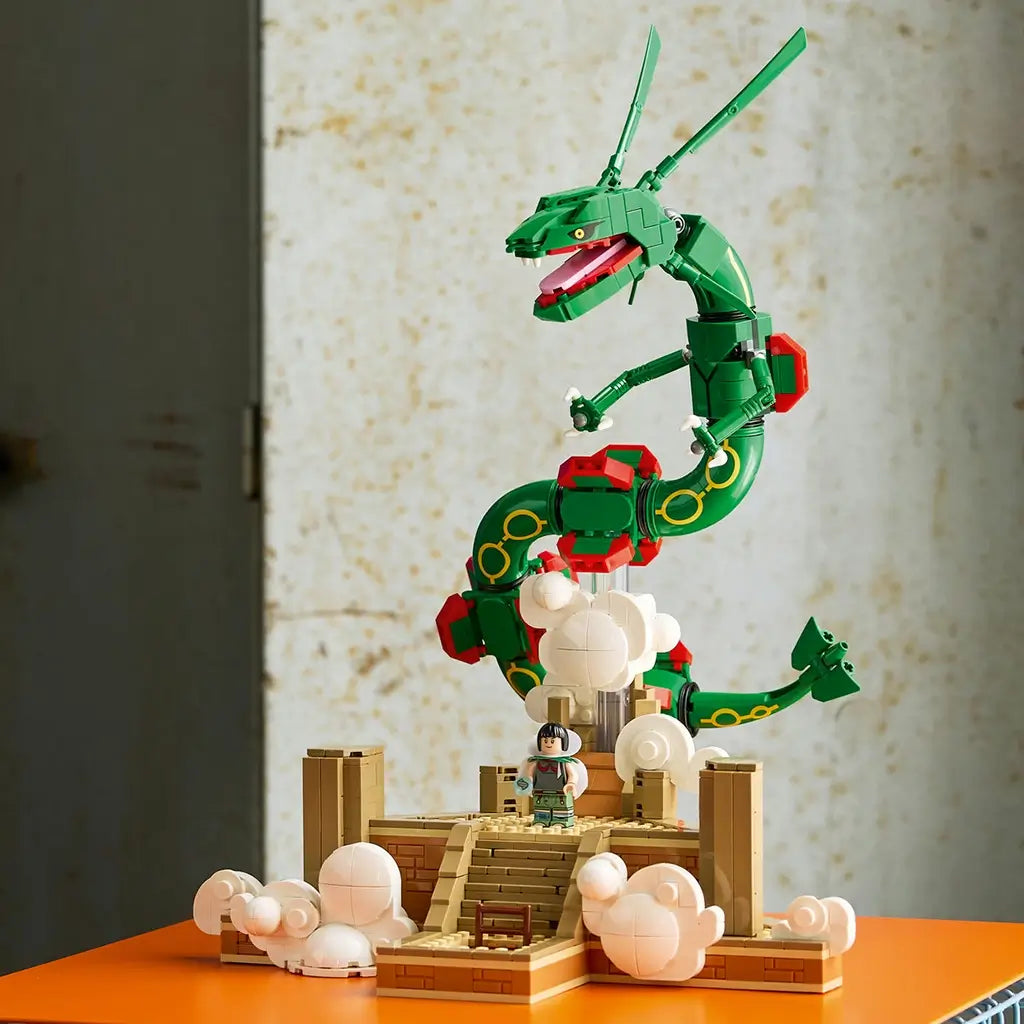 LEGO Pokémon Rayquaza display in green, red, tan, and white bricks features the articulated dragon above a cloud-covered shrine and trainer minifigure on an orange surface.