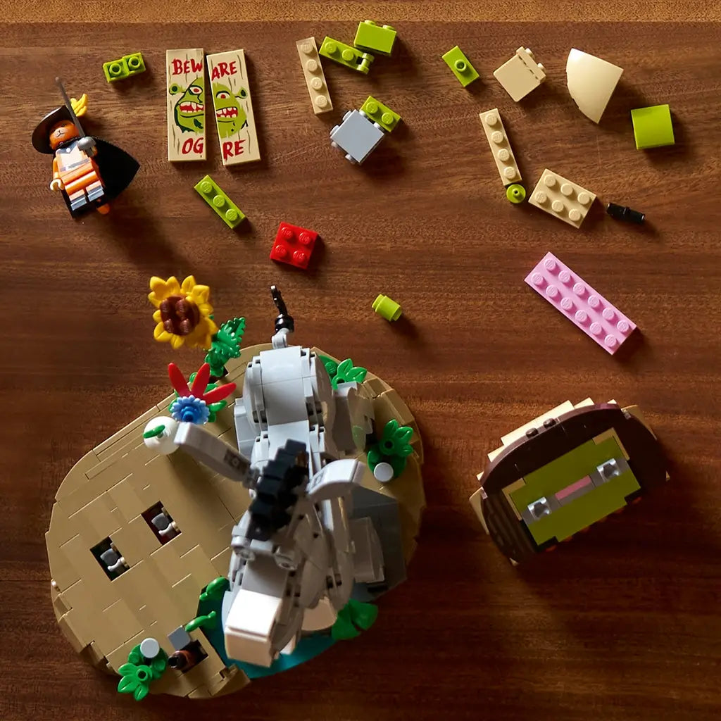 LEGO Shrek building set is shown partly assembled from above on a wooden table, with gray Donkey, orange Puss in Boots, colorful bricks, flowers and a Beware Ogre sign.
