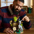 LEGO Shrek display model shows an adult adjusting gray Donkey beside green Shrek and orange Puss in Boots, with a Beware Ogre sign and plants on a wooden table.
