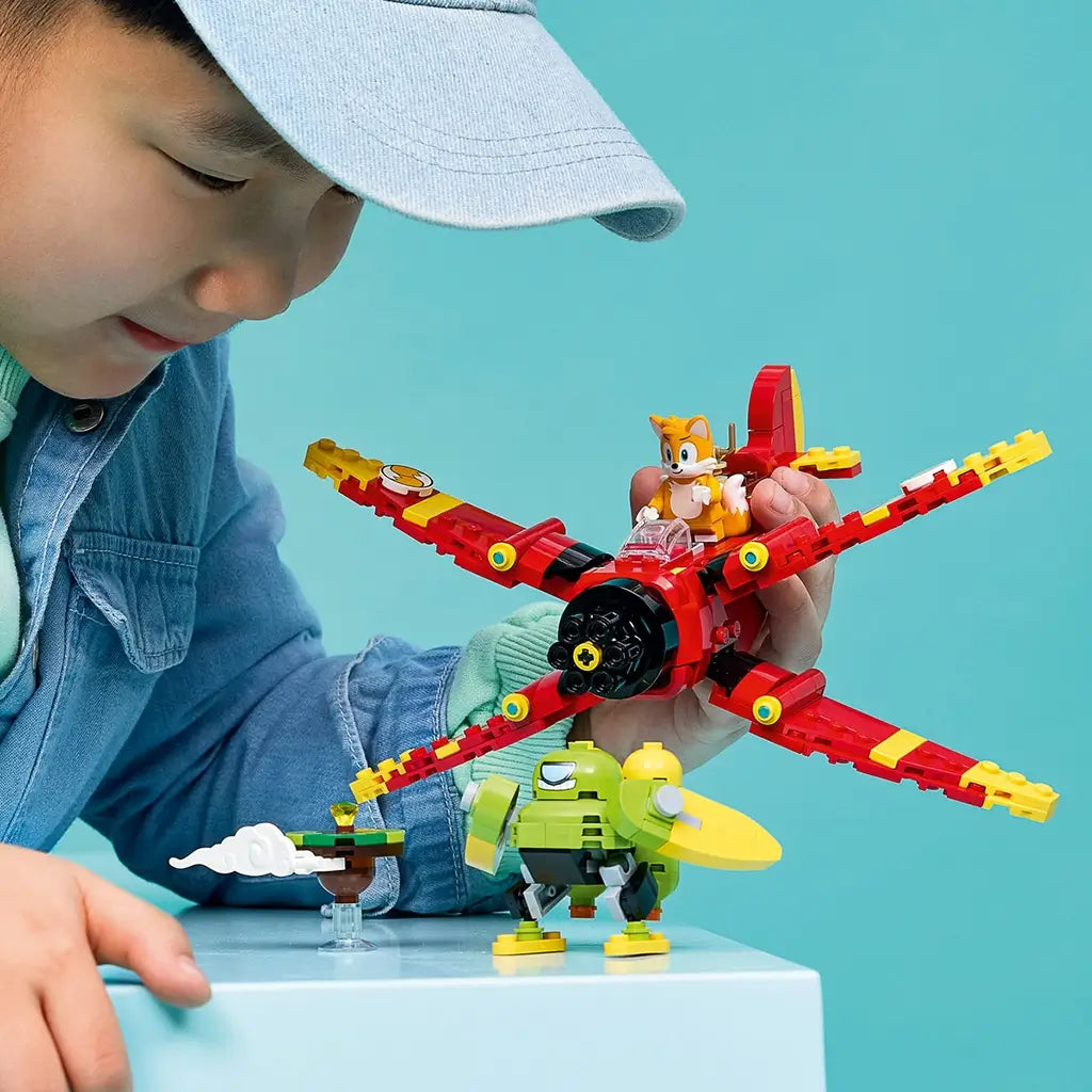 LEGO Sonic the Hedgehog Tails' Tornado-1 play scene shows a child holding the red and yellow brick airplane with Tails above a green Badnik and accessory on a blue pedestal.