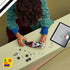 LEGO Audi F1 race car building scene in gray, black, and red plastic pieces, partially assembled model with scattered parts, child assembling at table with tablet instructions in background.