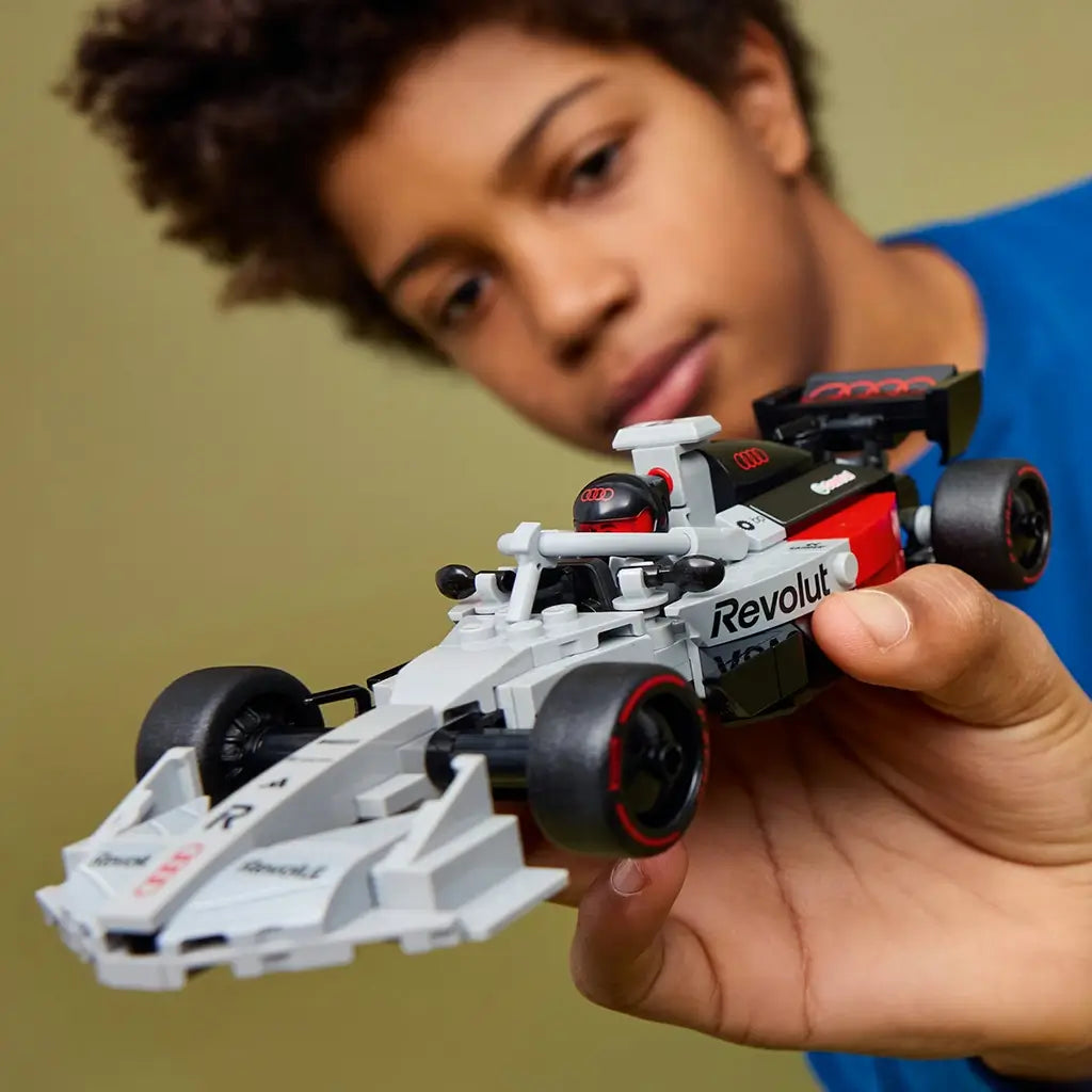 LEGO Audi F1 race car toy in gray, black, and red plastic, sleek Formula 1 shape, held by child for play, showcasing realistic wheels and cockpit detail against a soft neutral indoor background.