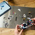 LEGO Speed Champions BMW M3 E30 build in white and black plastic sits partly assembled on a work surface with gold wheels, printed panels, a minifigure and tablet nearby.