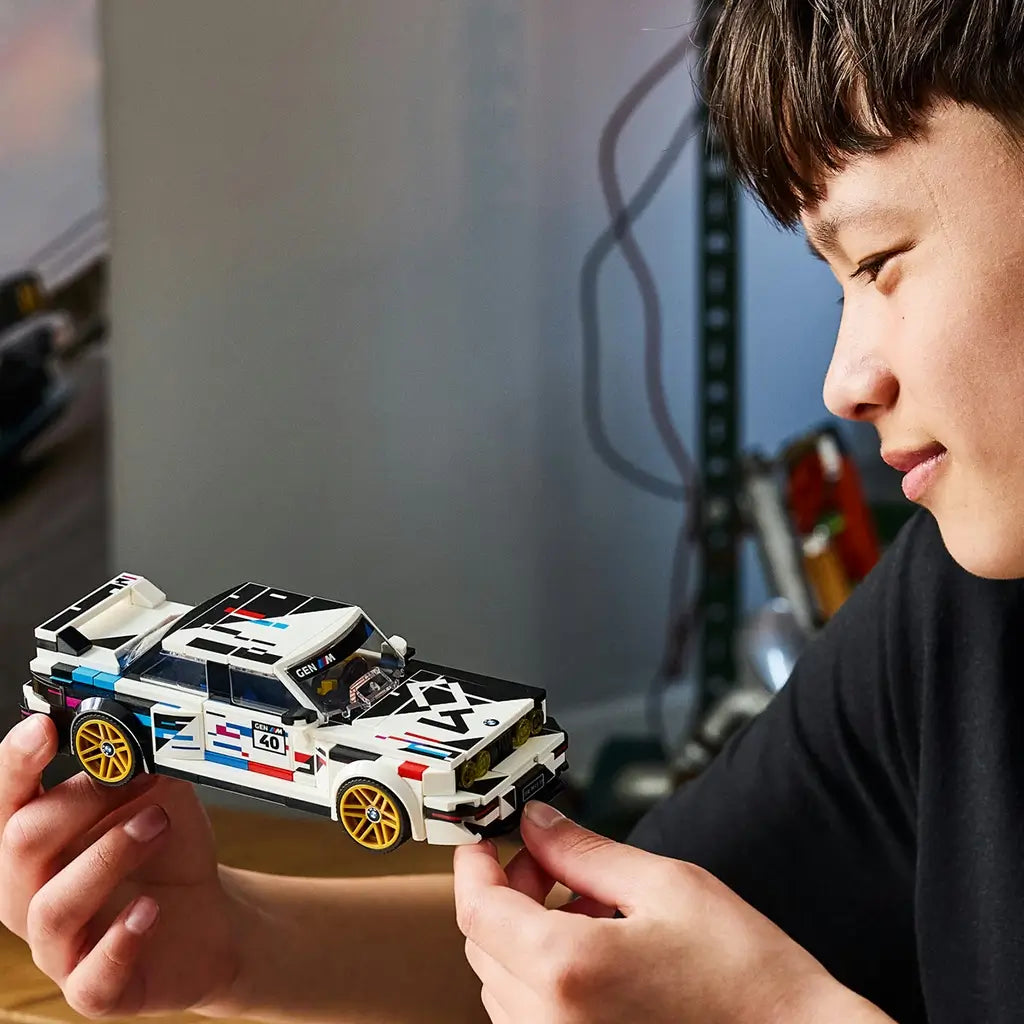 LEGO Speed Champions BMW M3 E30 in white and black plastic with gold wheels is held by a young builder, highlighting its angular body, racing graphics and rear spoiler.