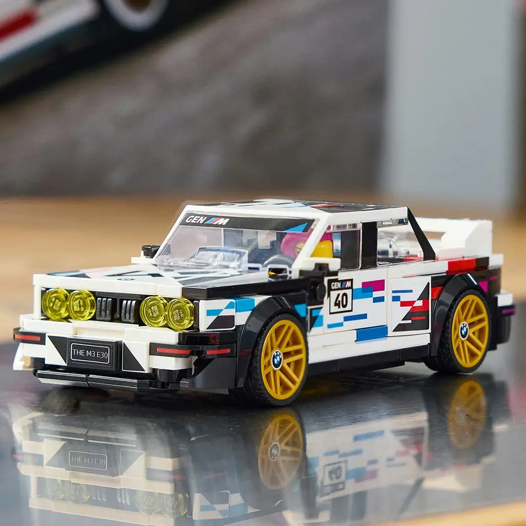 LEGO Speed Champions BMW M3 E30 in white and black plastic features gold wheels, yellow headlights and colorful racing graphics, displayed on a reflective indoor surface.
