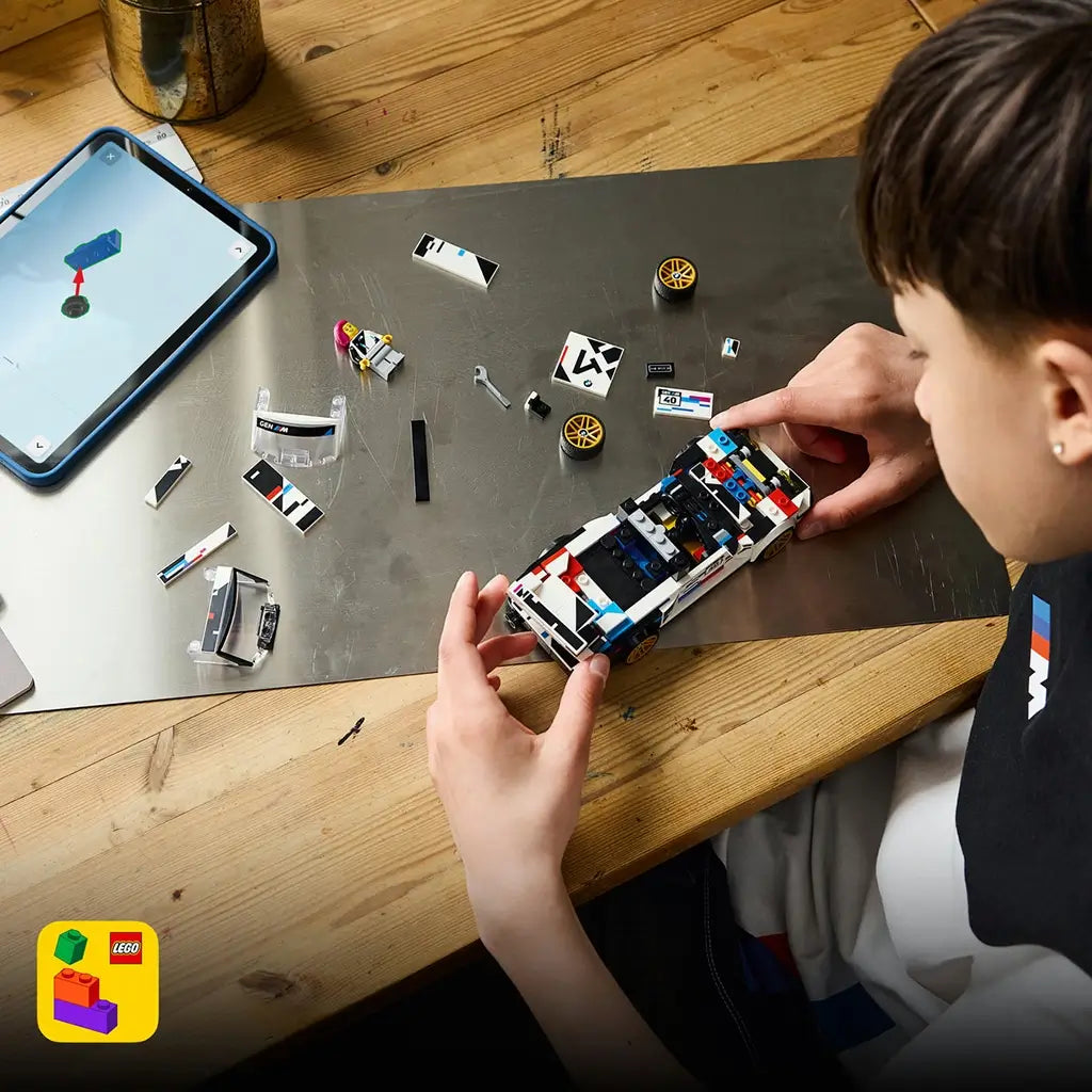 LEGO Speed Champions BMW M3 E30 in white and black plastic is assembled by a young builder at a wooden table with loose car pieces, gold wheels and a tablet nearby.