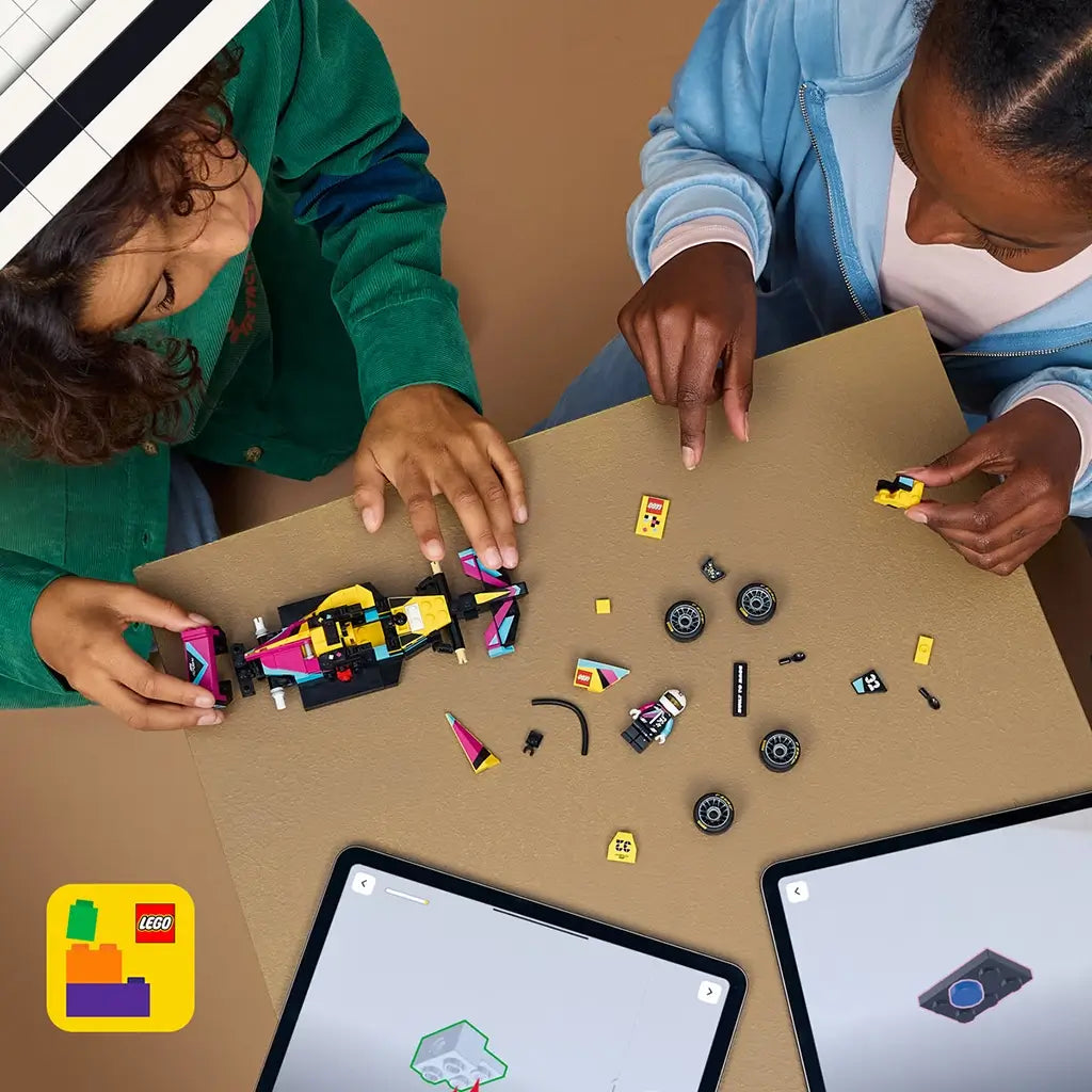 LEGO F1 Academy race car set, yellow black pink and teal plastic building toy pieces arranged during assembly, with tablets showing digital instructions on a tan tabletop as builders collaborate.