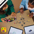 LEGO F1 Academy race car set, yellow black pink and teal plastic building toy pieces arranged during assembly, with tablets showing digital instructions on a tan tabletop as builders collaborate.