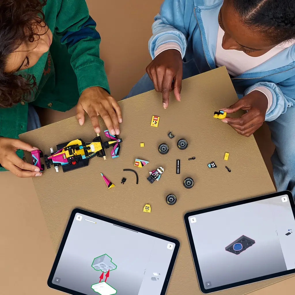 LEGO building experience, colorful plastic F1 Academy race car components and minifigure parts arranged on a tabletop, with two builders using digital instructions on tablets during assembly.