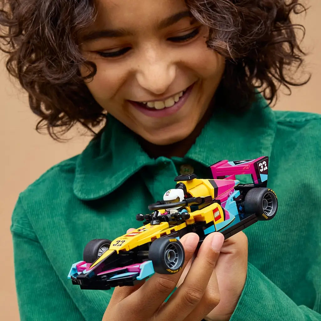 LEGO F1 Academy race car, yellow black pink and teal plastic Formula 1 model with aerodynamic shape, held by a smiling builder in a green jacket against a warm tan background.