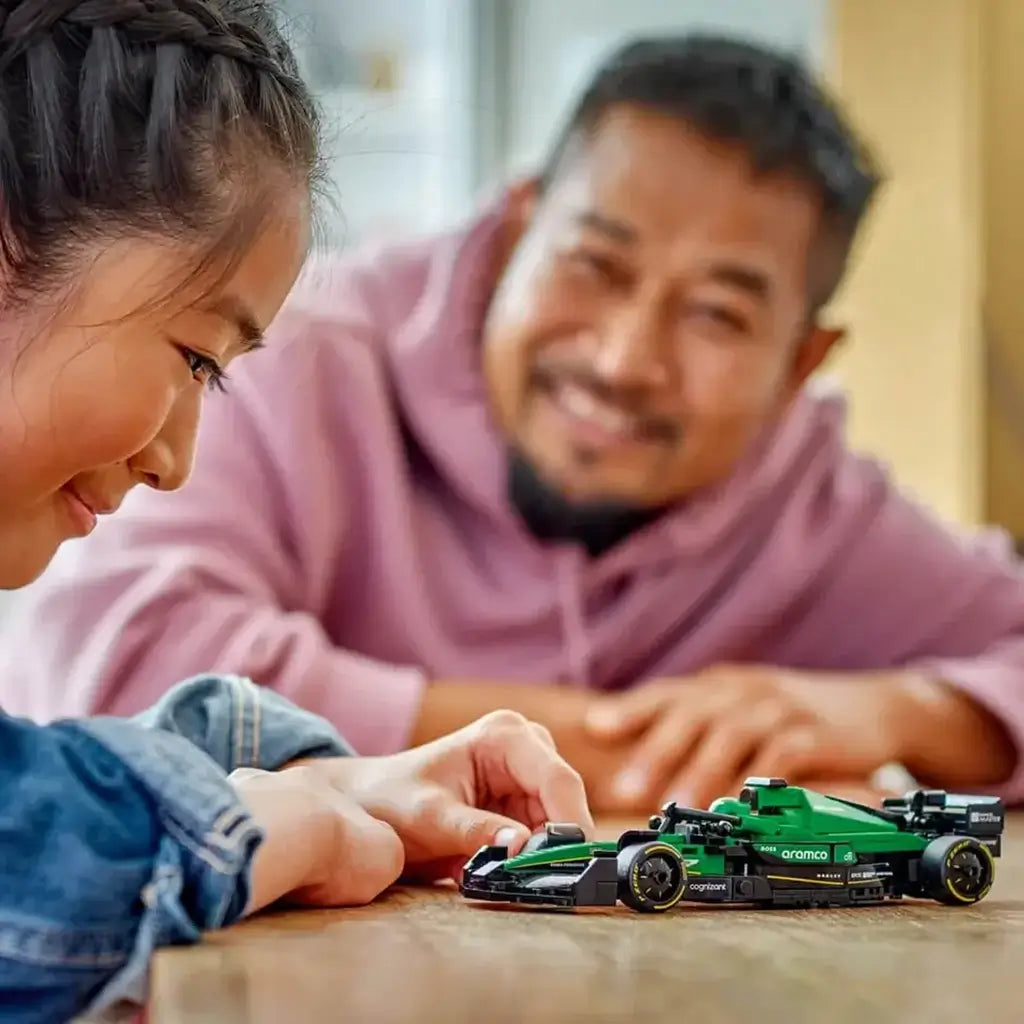 LEGO Speed Champions Aston Martin Aramco F1 model in green and black plastic with streamlined race car styling as a child plays while an adult watches, on a wooden tabletop indoors.