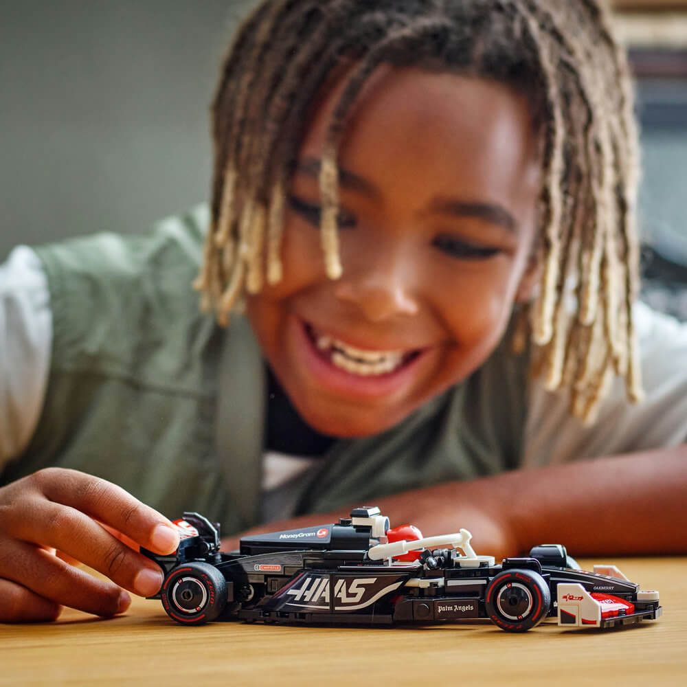 LEGO Speed Champions MoneyGram Haas F1 Team VF-24 black and red race car is pushed by a smiling child across a wooden tabletop during imaginative indoor play with authentic sponsor graphics.