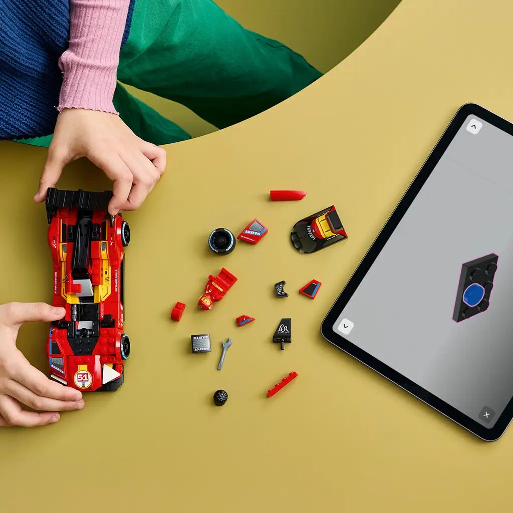 LEGO Speed Champions Ferrari 499P in red plastic is assembled on a yellow table with loose car parts and a tablet displaying digital building instructions beside it.