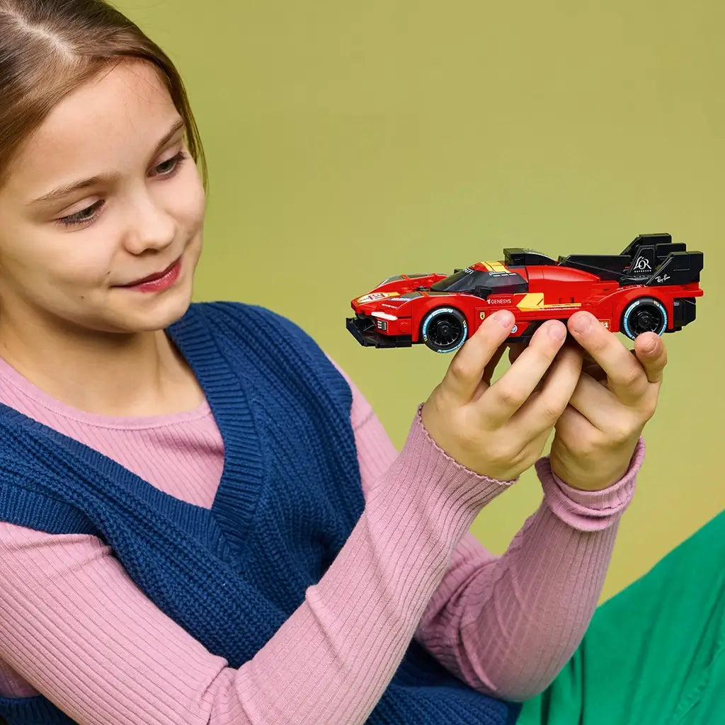 LEGO Speed Champions Ferrari 499P in red plastic with black aero details is held by a young builder, showing its low race-car shape and rear wing against a green backdrop.
