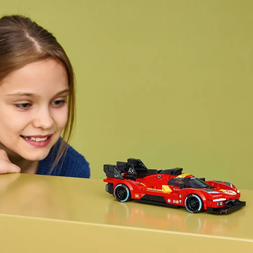 LEGO Speed Champions Ferrari 499P in red plastic with black wheels and racing decals sits on a yellow tabletop as a young builder looks closely at the finished model.