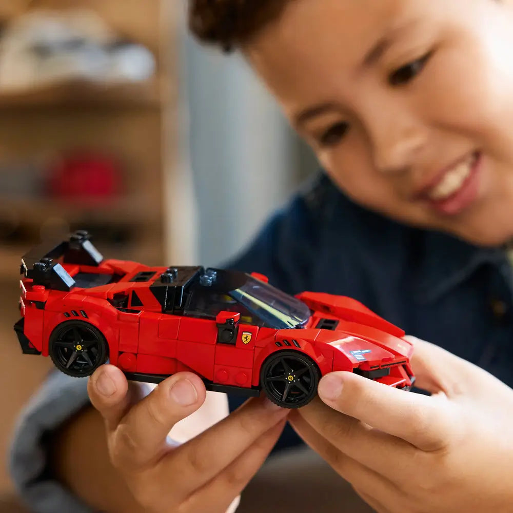 Young builder smiling while holding the completed LEGO Ferrari SF90 XX Stradale, highlighting the red bodywork, black wheels, rear wing, and detailed cockpit in a bright indoor setting