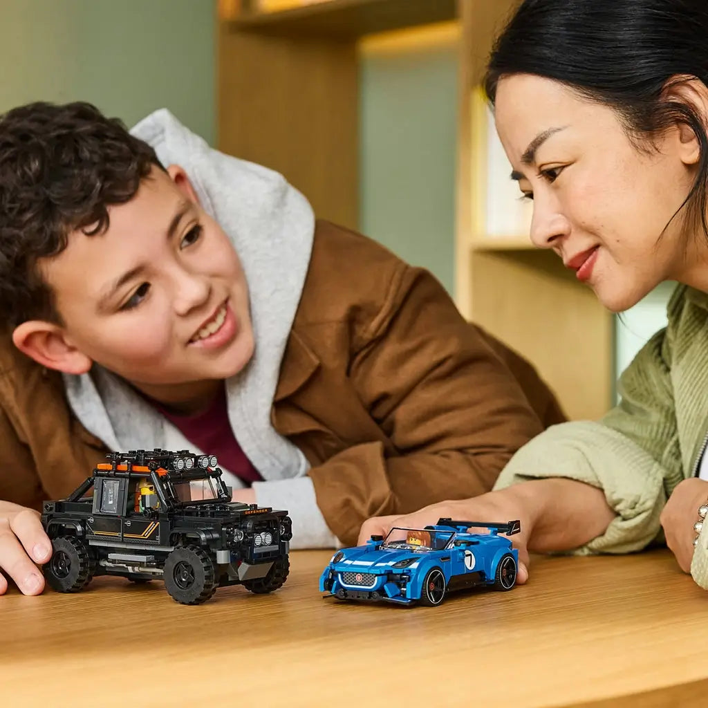 LEGO Speed Champions Jaguar and Land Rover models in blue and black plastic sit on a wooden table as a child and adult play with the brick-built vehicles together indoors.