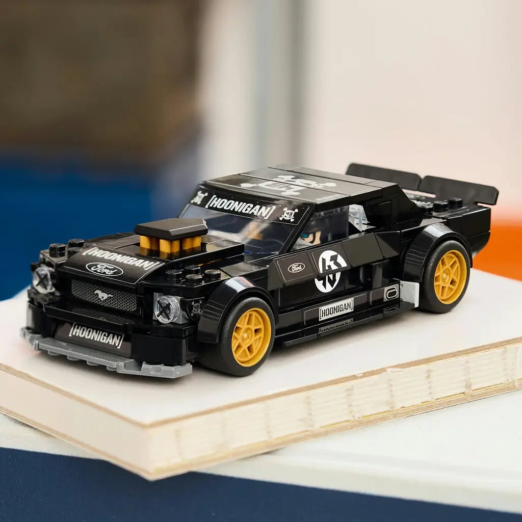 LEGO Speed Champions Ford Mustang Hoonicorn in glossy black plastic features gold wheels, Hoonigan graphics and a rear spoiler, displayed on a book in a softly lit room.