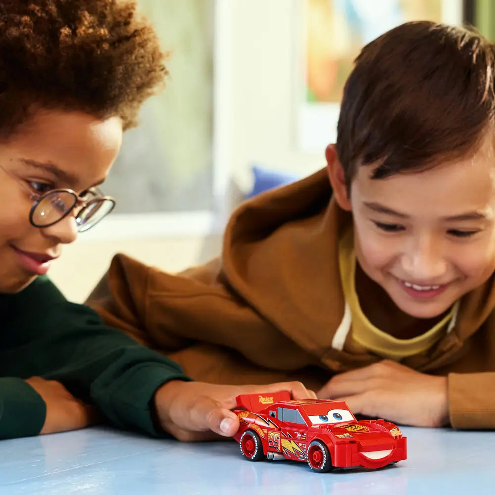 Two children smiling while playing with the completed LEGO Lightning McQueen car, showing the bright red build, expressive eyes, racing details, and a cheerful indoor play environment