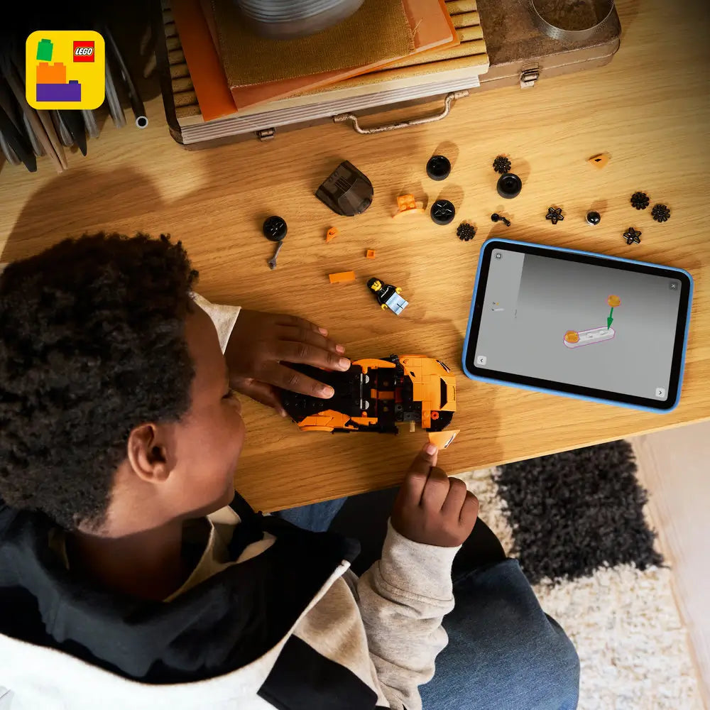 Child assembling the LEGO McLaren W1 supercar on a wooden desk, showing orange and black plastic car pieces, loose wheels, minifigure parts, and a tablet displaying step-by-step digital building instructions