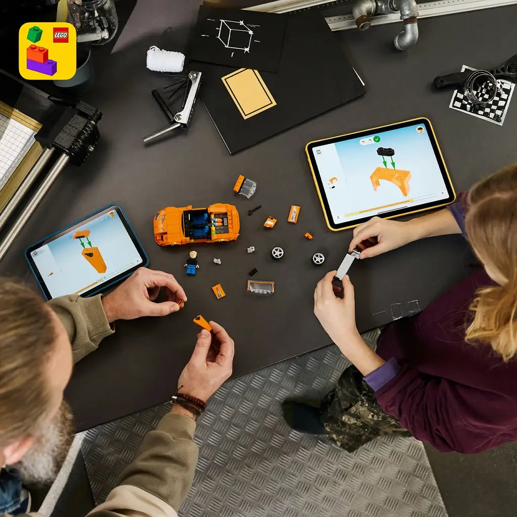 LEGO Speed Champions Toyota Supra MK4 build scene shows two people assembling the orange brick car with loose pieces and tablets displaying digital instructions on a dark workbench.