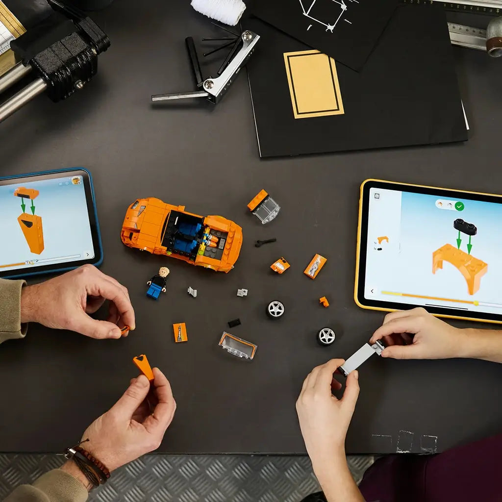 LEGO Speed Champions Toyota Supra MK4 in orange plastic is being assembled by two builders, with loose car parts and digital instructions shown on tablets across a dark worktable.