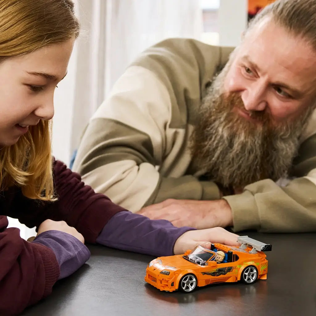 LEGO Speed Champions Toyota Supra MK4 in orange plastic sits on a dark tabletop as a young builder reaches toward the car while an adult watches from across the table.