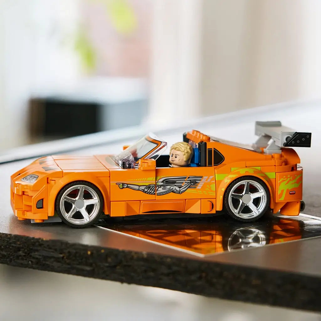 LEGO Speed Champions Toyota Supra MK4 in orange plastic features silver wheels, green side graphics and a gray rear wing, displayed in profile on a reflective indoor surface.
