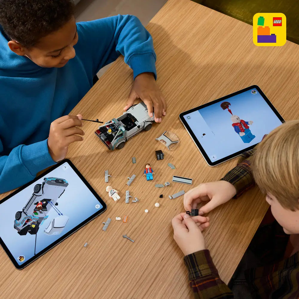 Two children building the LEGO Back to the Future Time Machine at a wooden table, with gray car parts, loose bricks, minifigures, and tablets showing step-by-step digital building instructions