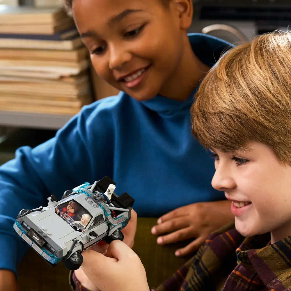 Two children smiling while holding the completed LEGO Back to the Future Time Machine, showing detailed interior, seated minifigures, and translucent blue energy elements in a cozy indoor setting
