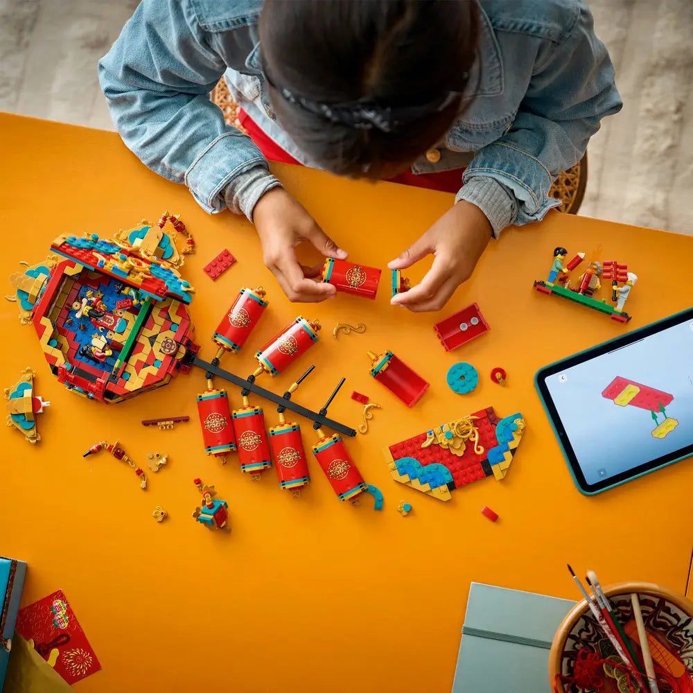 Child assembling the LEGO Fortune Firecracker set on a bright yellow table, with red firecracker elements, decorative panels, loose bricks, and a tablet displaying digital building instructions