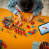Child assembling the LEGO Fortune Firecracker set on a bright yellow table, with red firecracker elements, decorative panels, loose bricks, and a tablet displaying digital building instructions