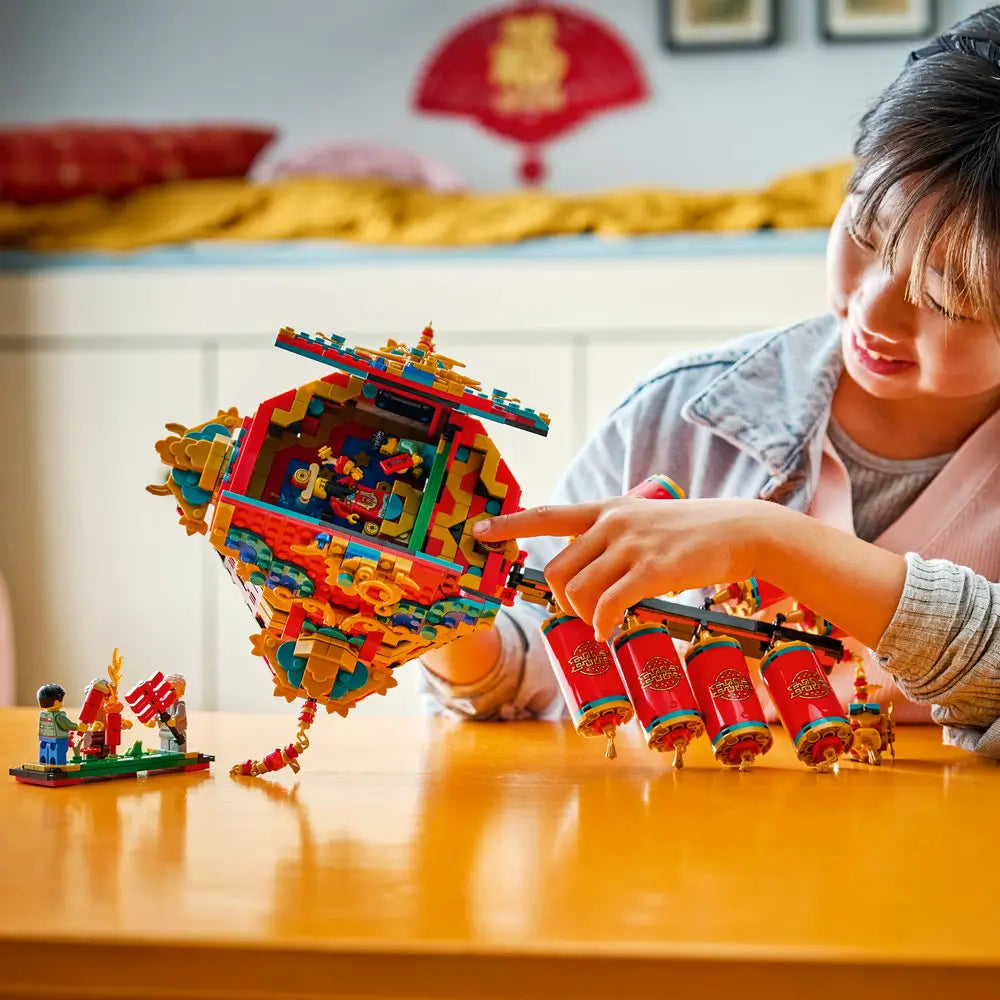Child holding and examining the LEGO Fortune Firecracker build, showing the opening decorative panels, hanging firecrackers, and detailed interior figures in a warm indoor setting