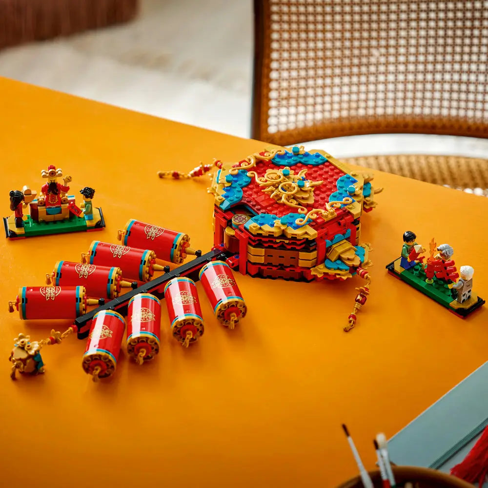 Completed LEGO Fortune Firecracker model displayed on a table, featuring red cylindrical firecrackers, gold accents, teal details, and Spring Festival minifig scenes on small bases