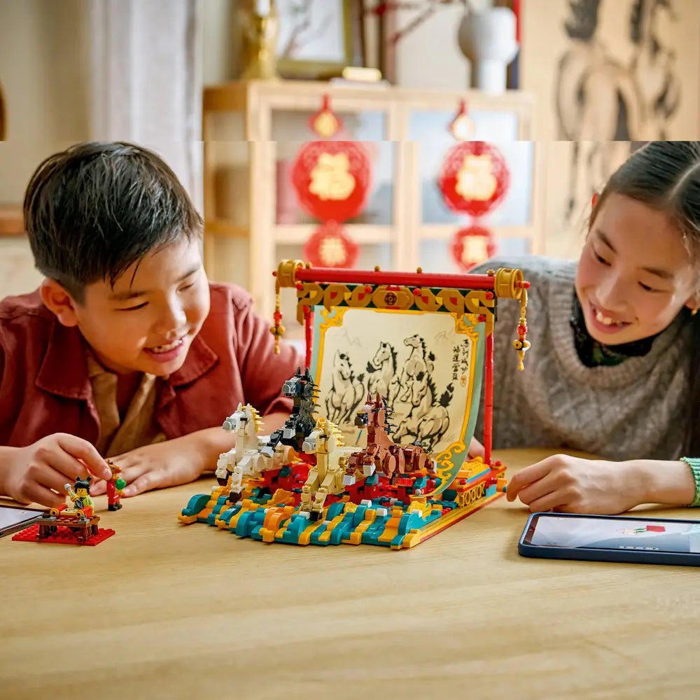Two children smiling as they assemble the LEGO Galloping Horses Canvas, with multicolored plastic bricks, horse figures, and festive red decorations displayed in a cozy home environment