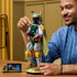 LEGO Star Wars Boba Fett figure in green, gray, red and yellow plastic bricks, with helmet and cape, adjusted by an adult builder beside a minifigure and display plaque.