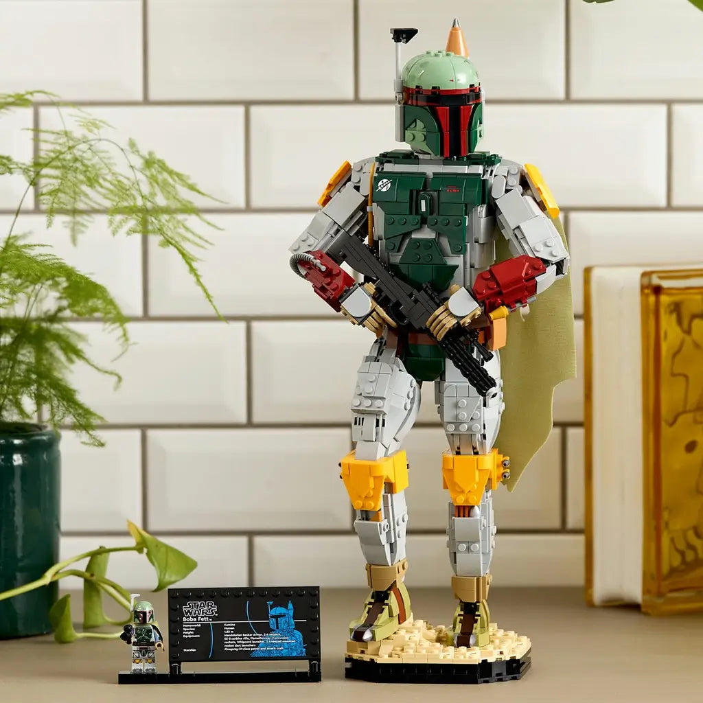 LEGO Star Wars Boba Fett display figure in green, gray, red and yellow plastic bricks, holding a black blaster beside a minifigure and plaque in a bright indoor setting.