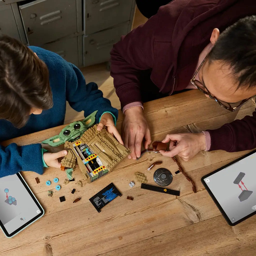 LEGO Star Wars Grogu build activity shows a green brick-built character being assembled with tan textured pieces, accessories, tablets and parts spread across a wooden table.