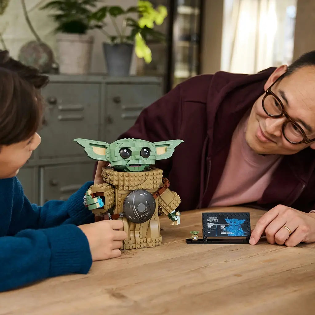 LEGO Star Wars Grogu building toy is shown with a child and adult, featuring a green brick-built character, tan textured robe and display plaque on a wooden tabletop indoors.