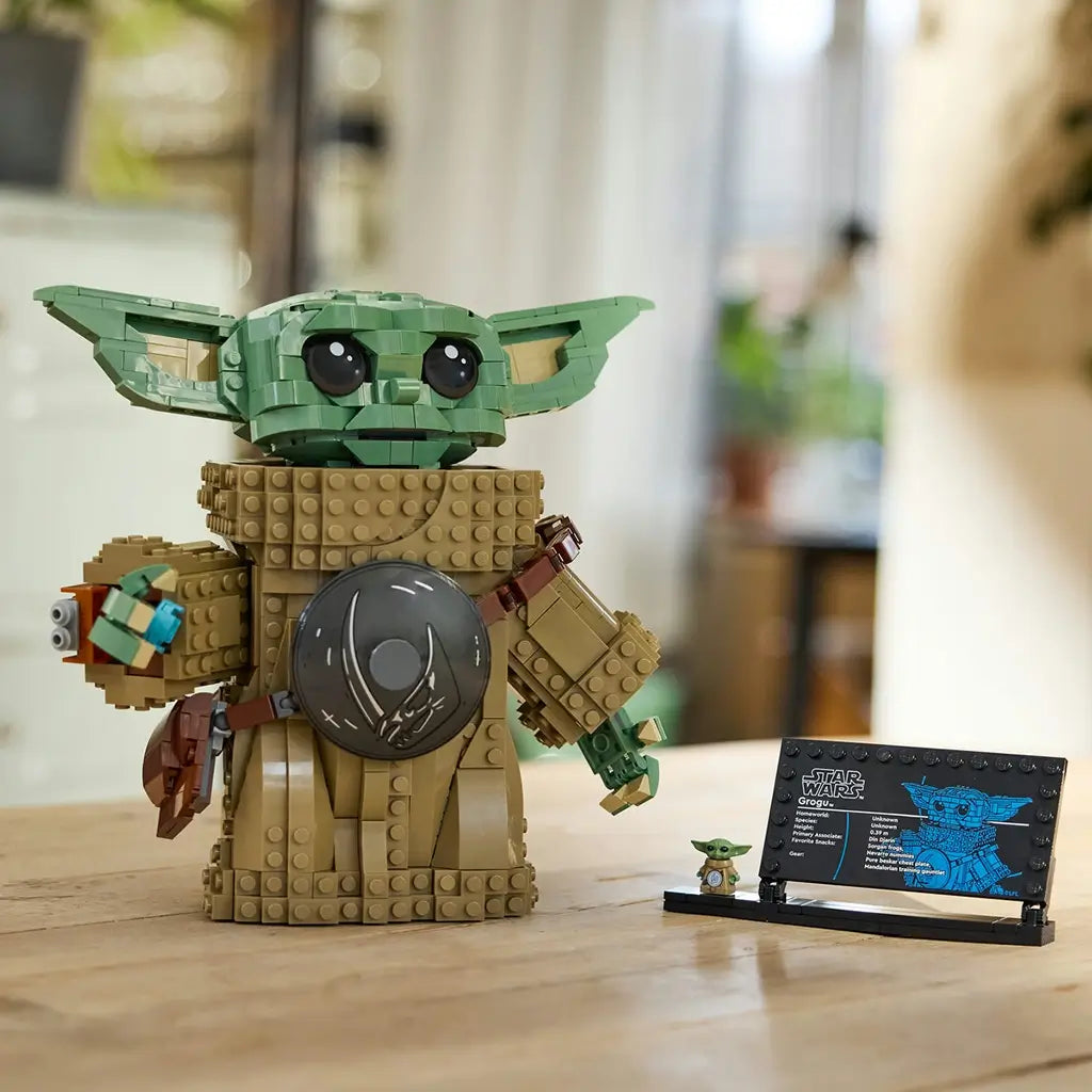 LEGO Star Wars Grogu model features a green brick-built head, tan textured robe and round silver accessory, displayed beside an information plaque on a wooden table in a home setting.