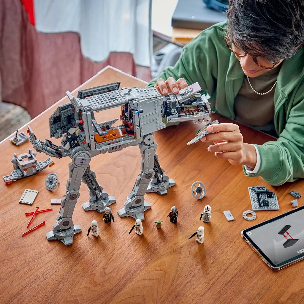 LEGO Star Wars Imperial Remnant AT-AT in gray plastic bricks is assembled by a builder at a wooden table, with open side panels, loose pieces, minifigures and a tablet.