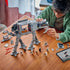 LEGO Star Wars Imperial Remnant AT-AT in gray plastic bricks is assembled by a builder at a wooden table, with open side panels, loose pieces, minifigures and a tablet.