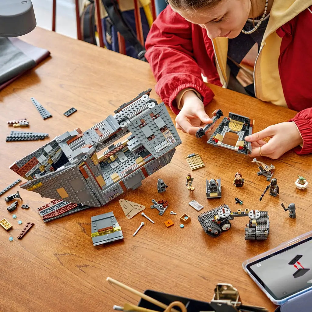 LEGO Star Wars Offworld Sandcrawler build in gray, tan and brown plastic is partly assembled across a wooden table as a young builder adds pieces beside minifigures and a tablet.