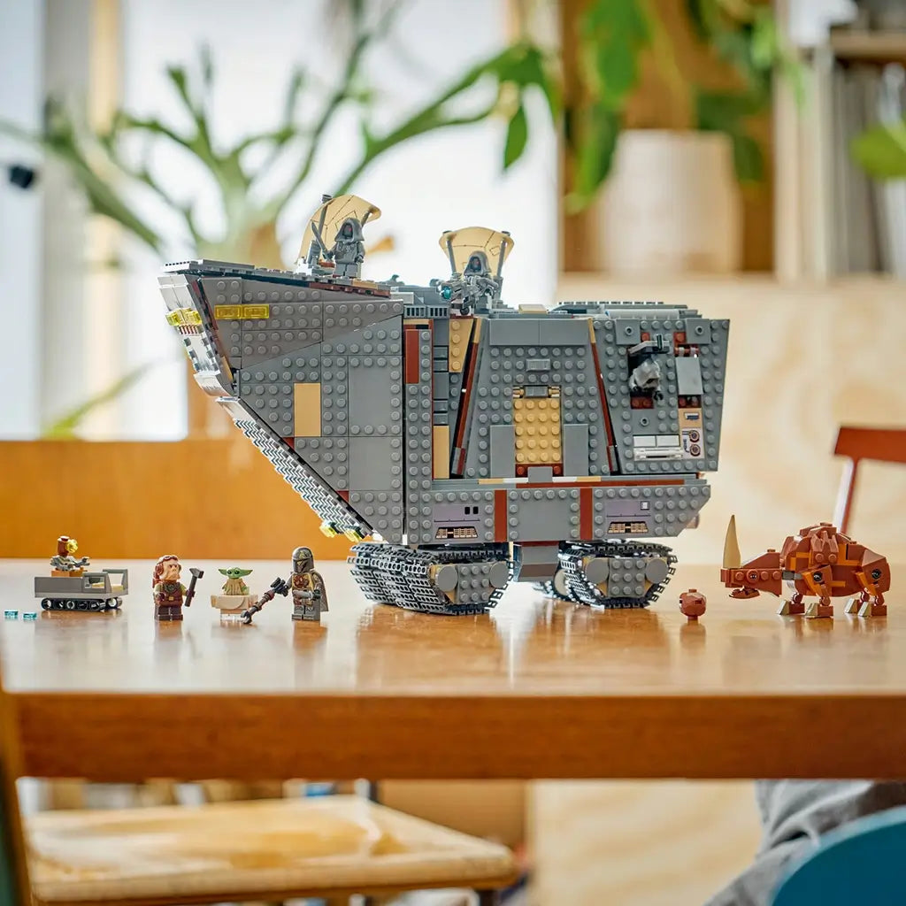 LEGO Star Wars Offworld Sandcrawler in gray plastic stands on black tracks with minifigures, Grogu, a cart and brown Mudhorn displayed across a wooden table in a bright room.