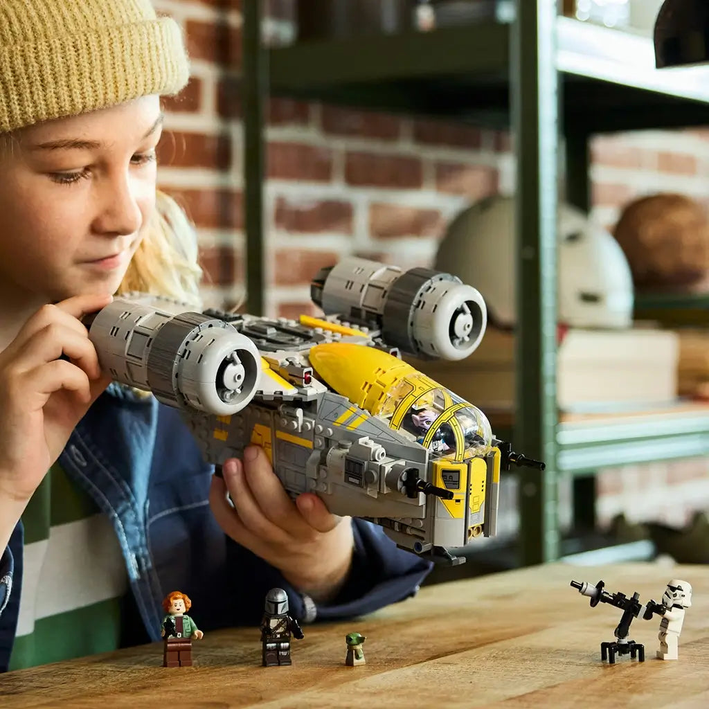 LEGO Star Wars Razor Crest vehicle, gray and yellow plastic starship with broad fuselage, twin engines and action-play theme, held by a builder in a workshop setting with minifigures.