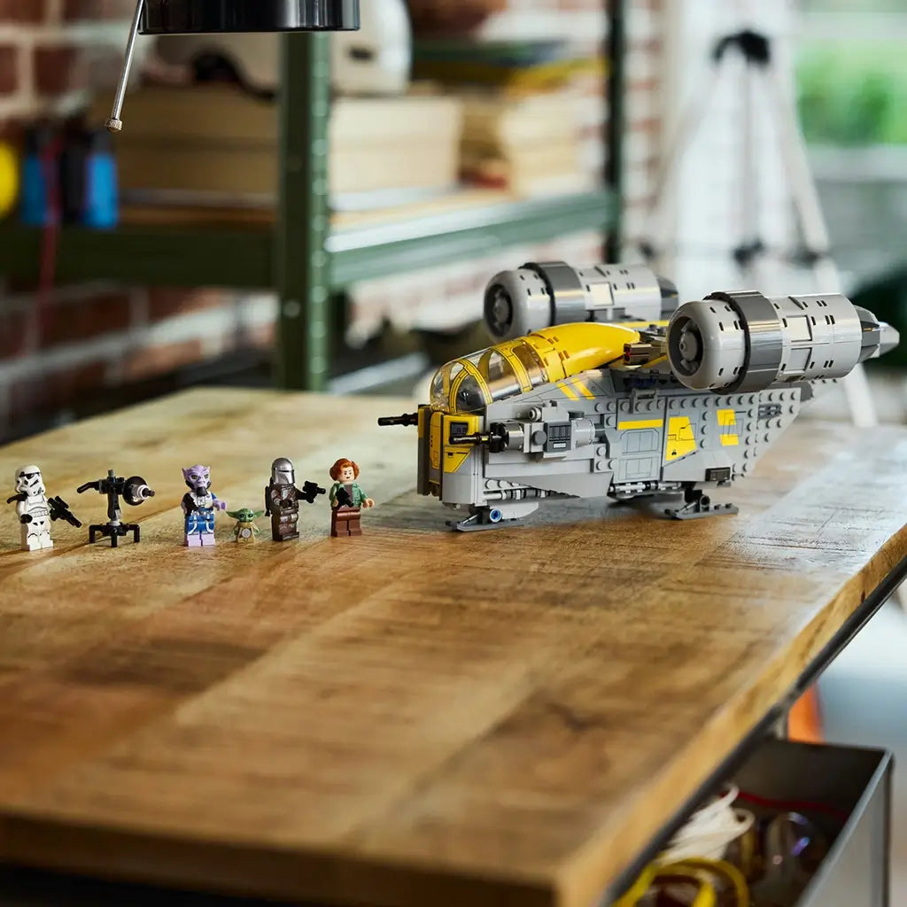 LEGO Star Wars Razor Crest model, gray and yellow plastic starship with curved cockpit canopy, twin engines and display-ready form, arranged with minifigures on a workshop tabletop background.